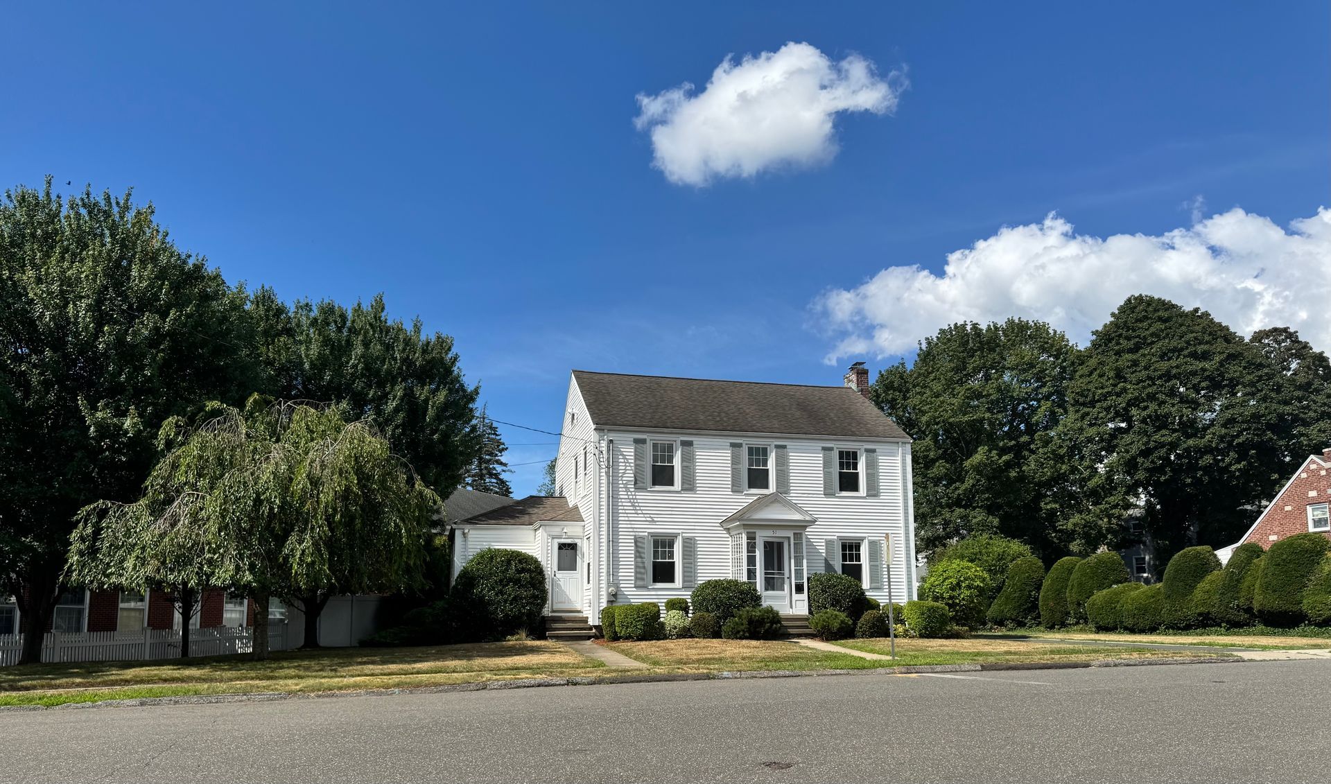 White two-story house with shutters, surrounded by trees, under a bright blue sky with clouds.