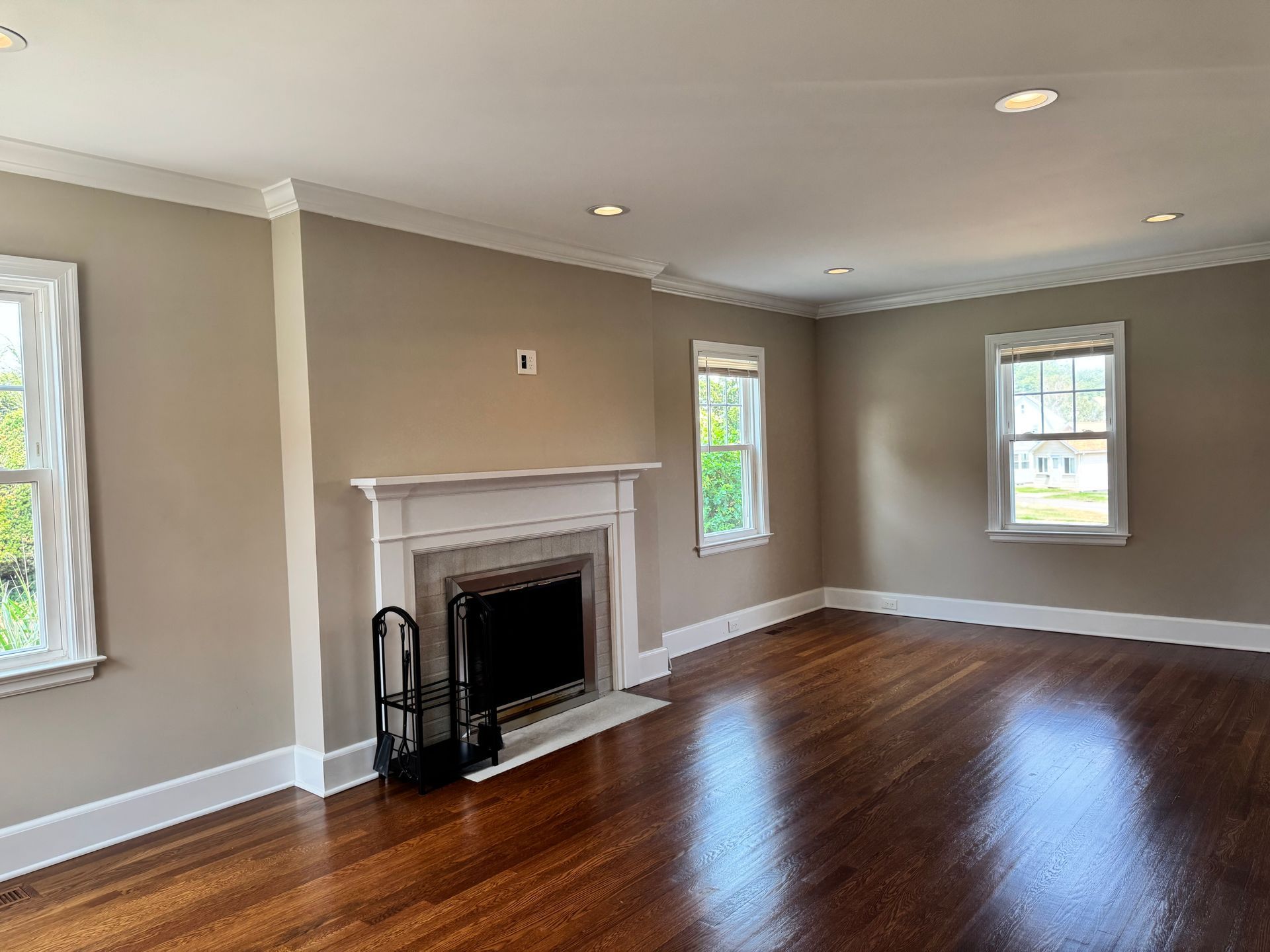 Empty living room with fireplace, hardwood floor, beige walls, white trim, two windows.