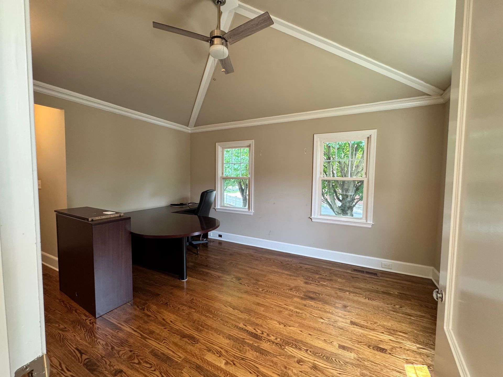 Empty office with brown desk and hardwood floors, two windows, and vaulted ceiling.