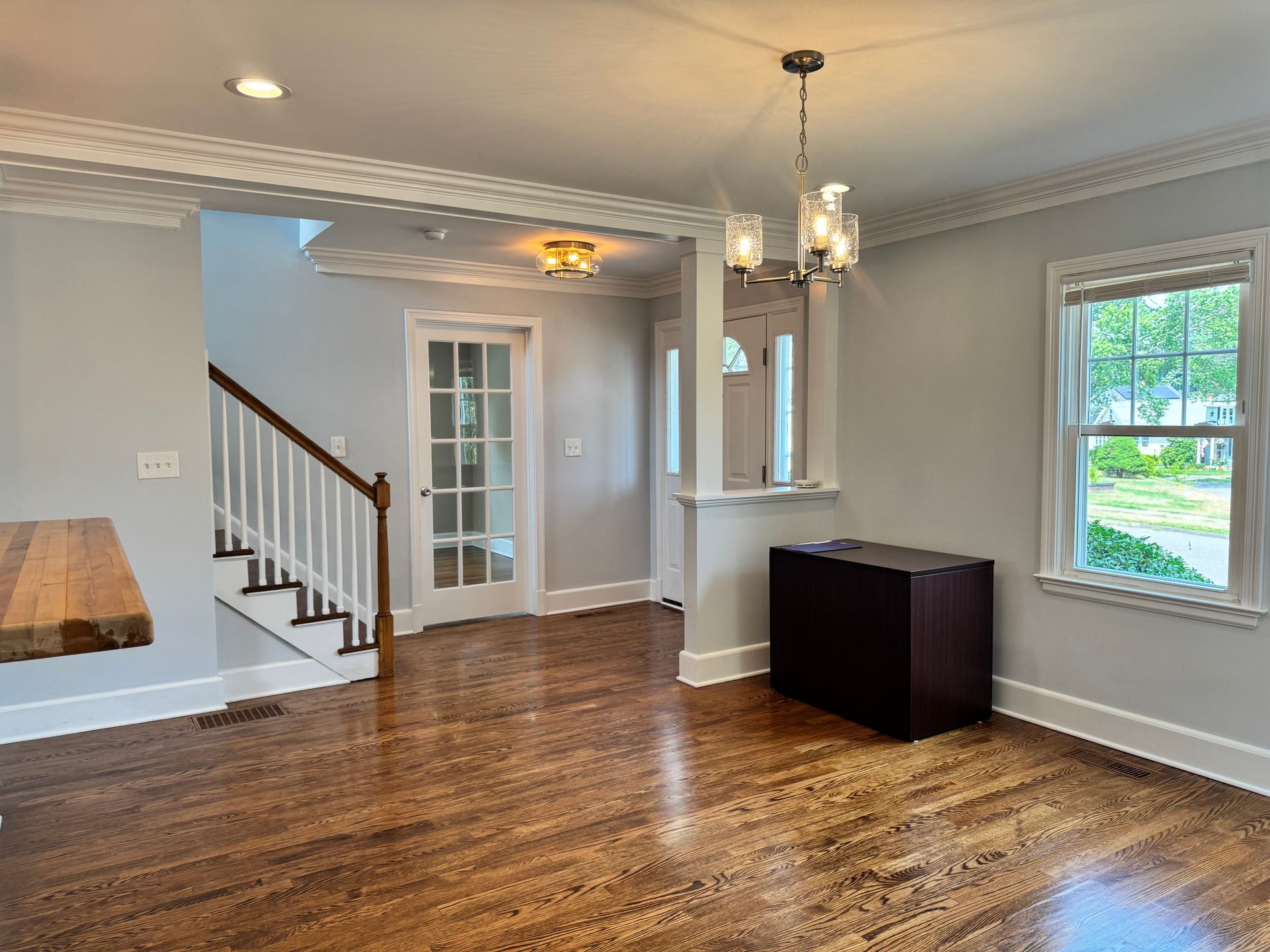 Interior view of a room with hardwood floors, stairs, chandelier, and a dark cabinet. Gray walls and white trim.