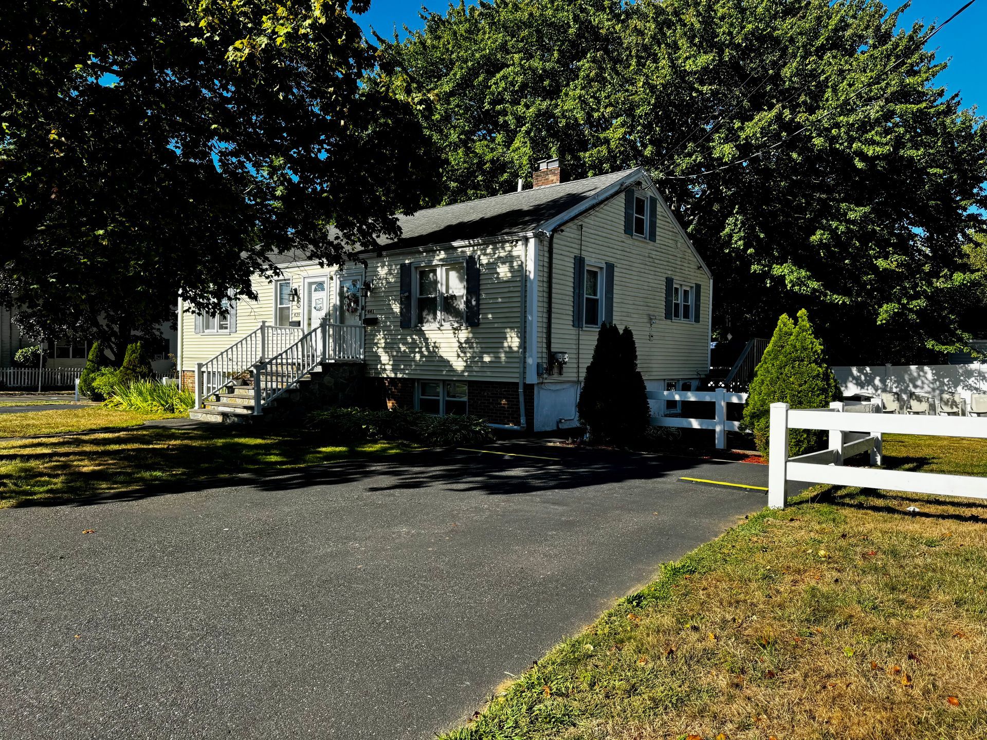 Beige two-story house with dark driveway, white fence, and green trees.