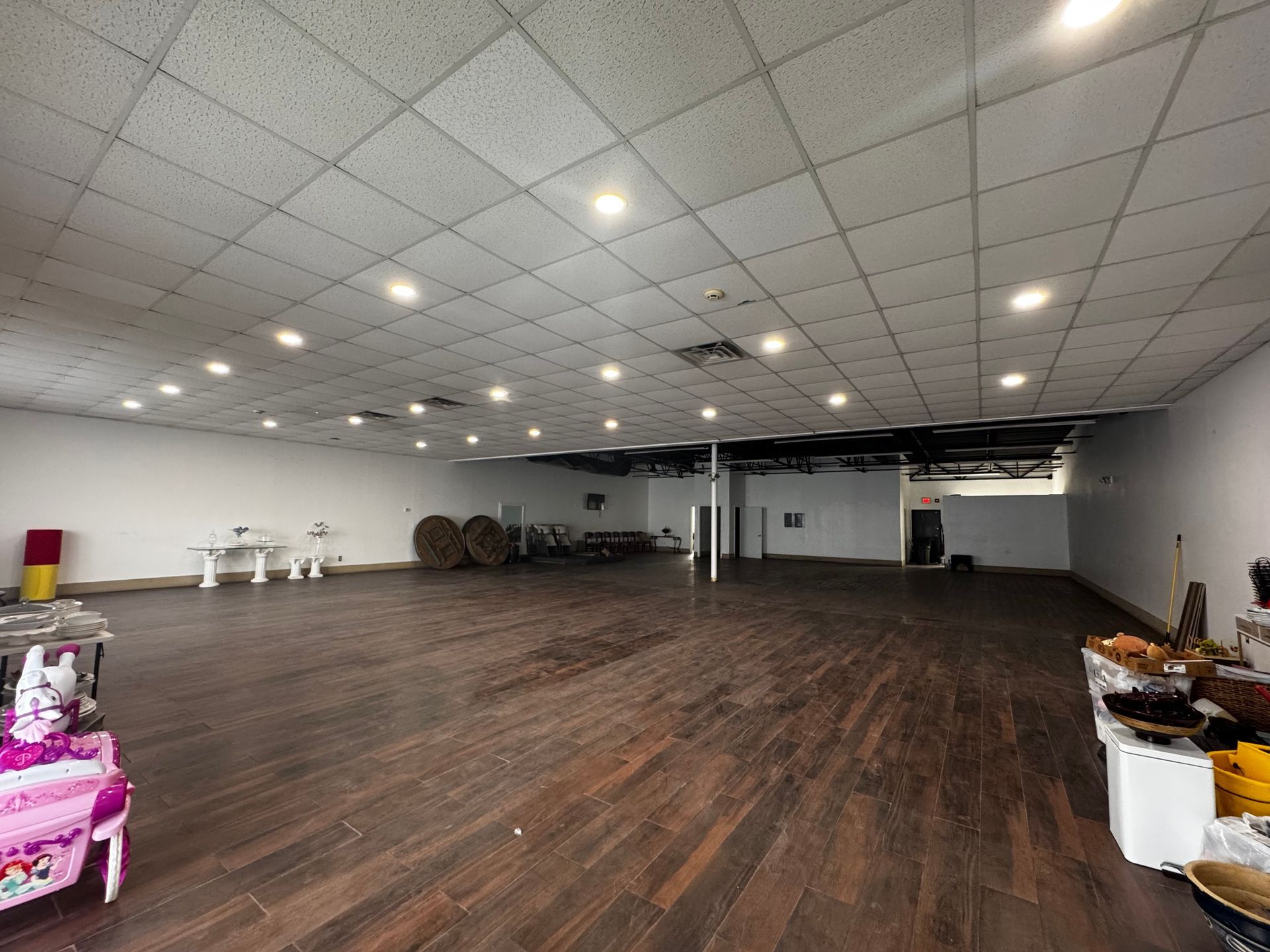 Empty, large room with wooden floor, white walls, and ceiling lights. Some items are placed along the side.