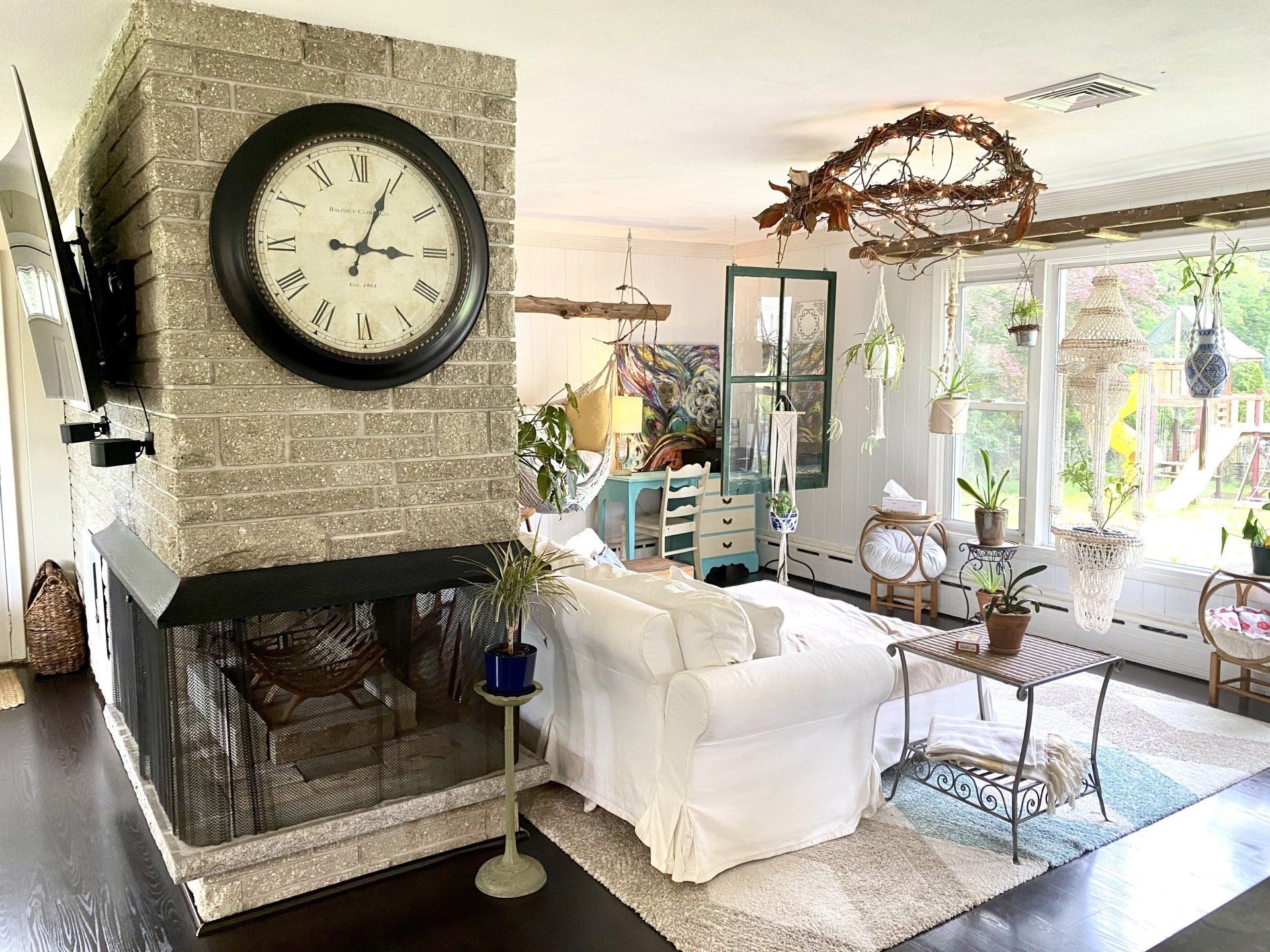 Living room with large clock on fireplace, white sofa, plants, and natural light.