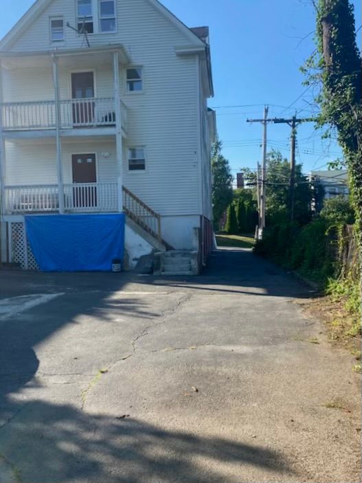 Back view of a white multi-story building with a blue tarp. Narrow asphalt alleyway leads away.