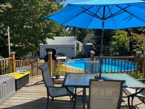 Wooden deck with blue umbrella, table, chairs, and above-ground pool in a backyard setting.