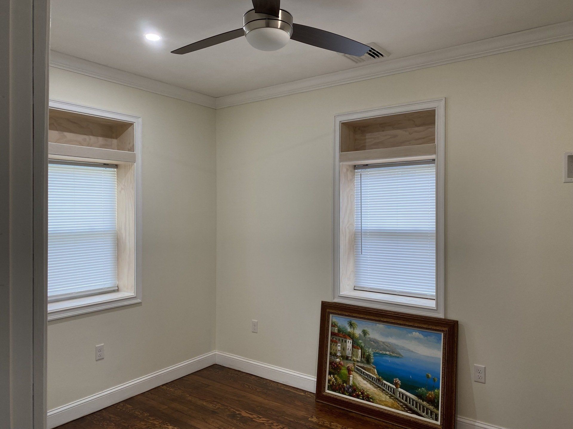 Empty room with two windows, ceiling fan, and framed artwork.