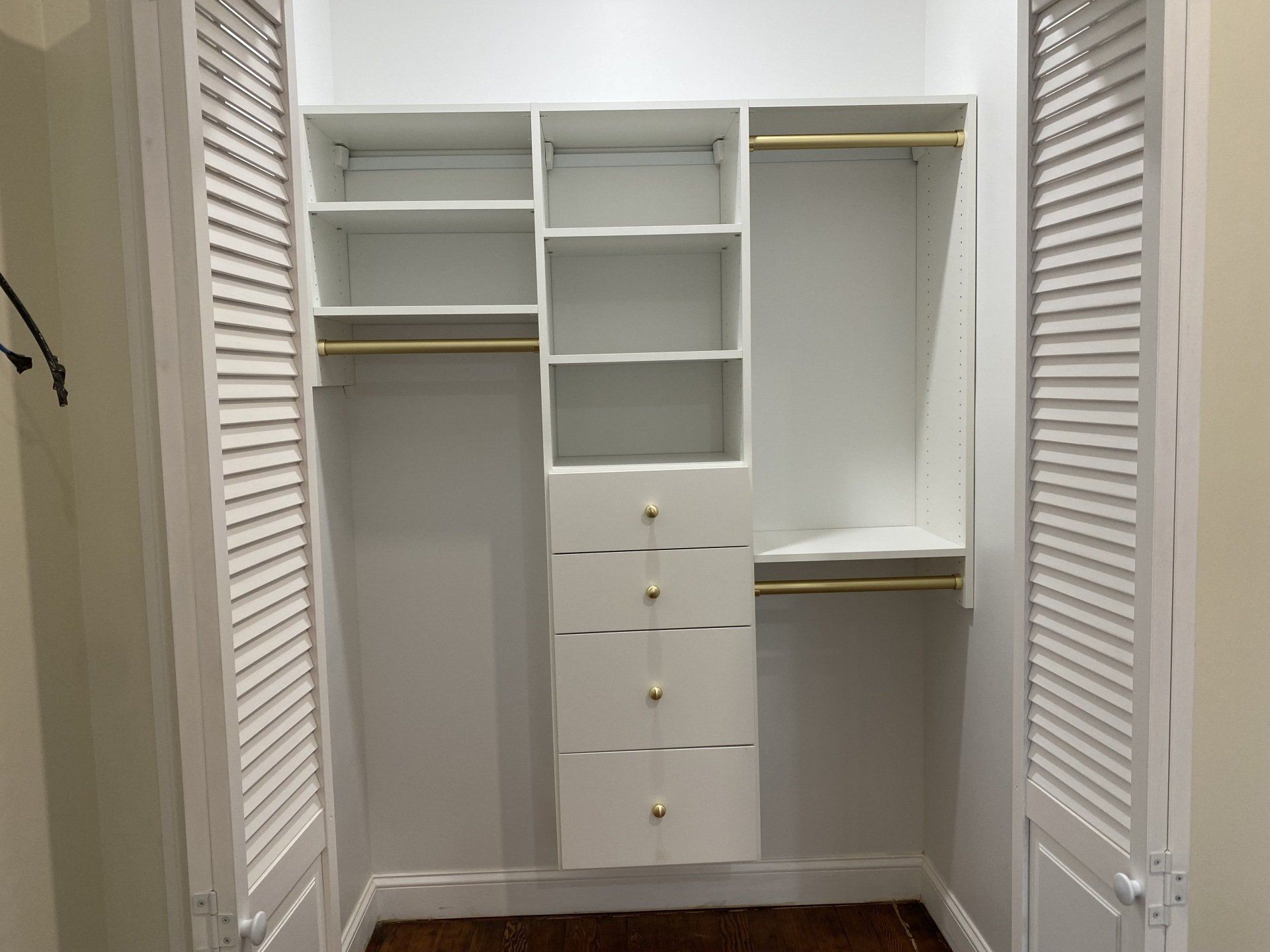 White closet organizer with shelves, drawers, and gold rods. Shutter-style doors open.