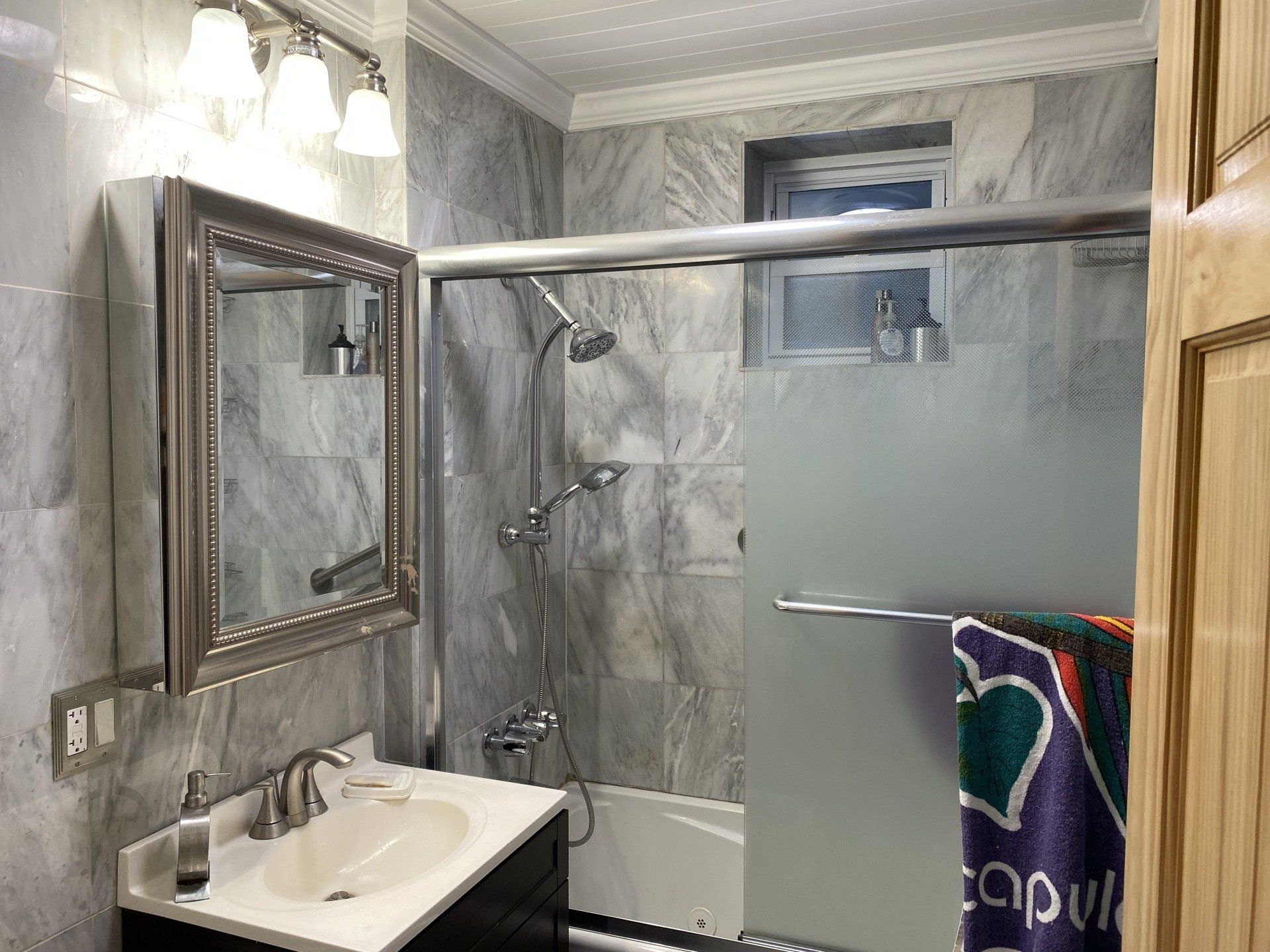 Bathroom with grey tile walls, shower, vanity, and mirror with a light fixture.