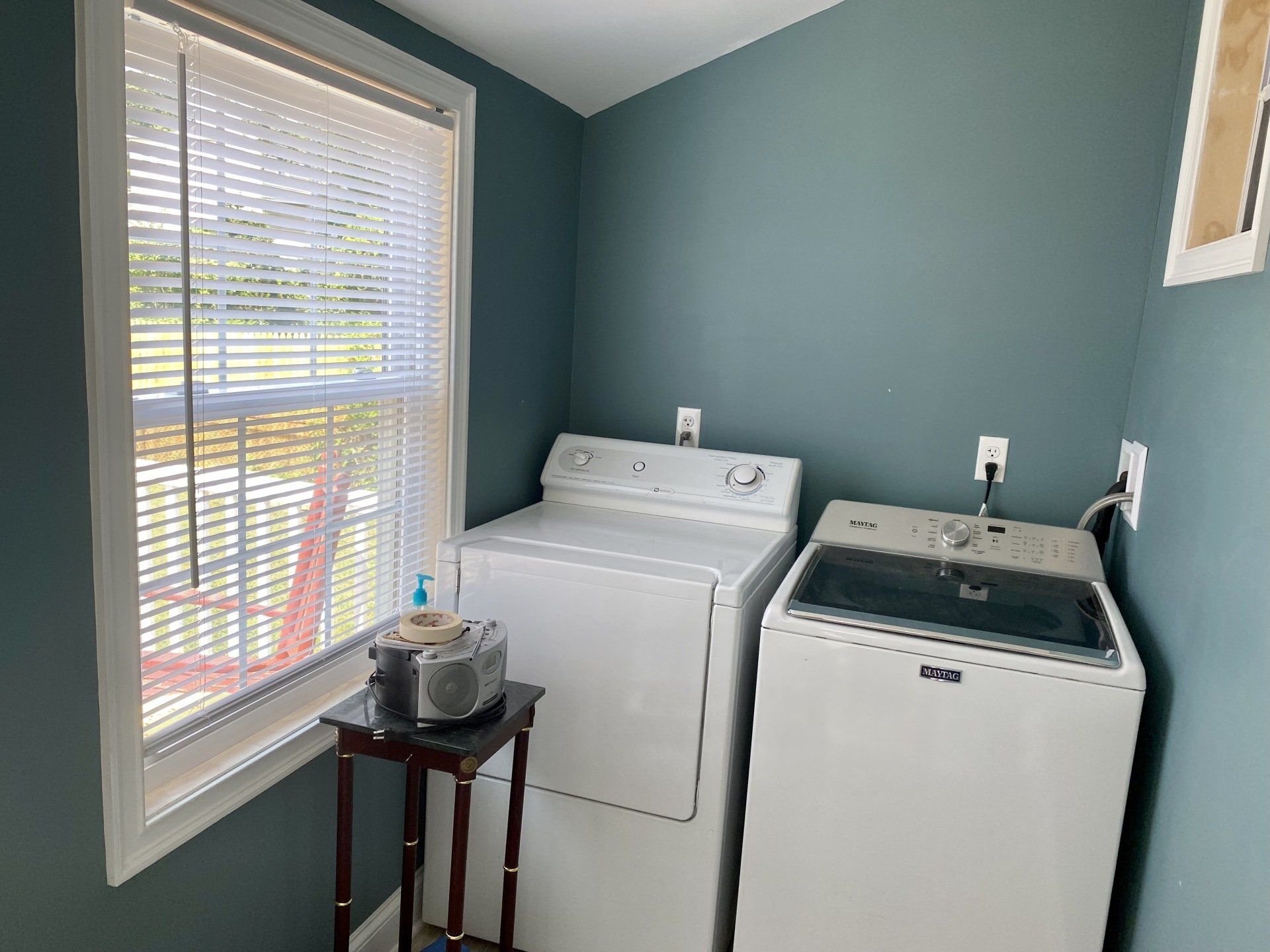 Laundry room with white washer and dryer, blue walls, window with blinds, small table with items.
