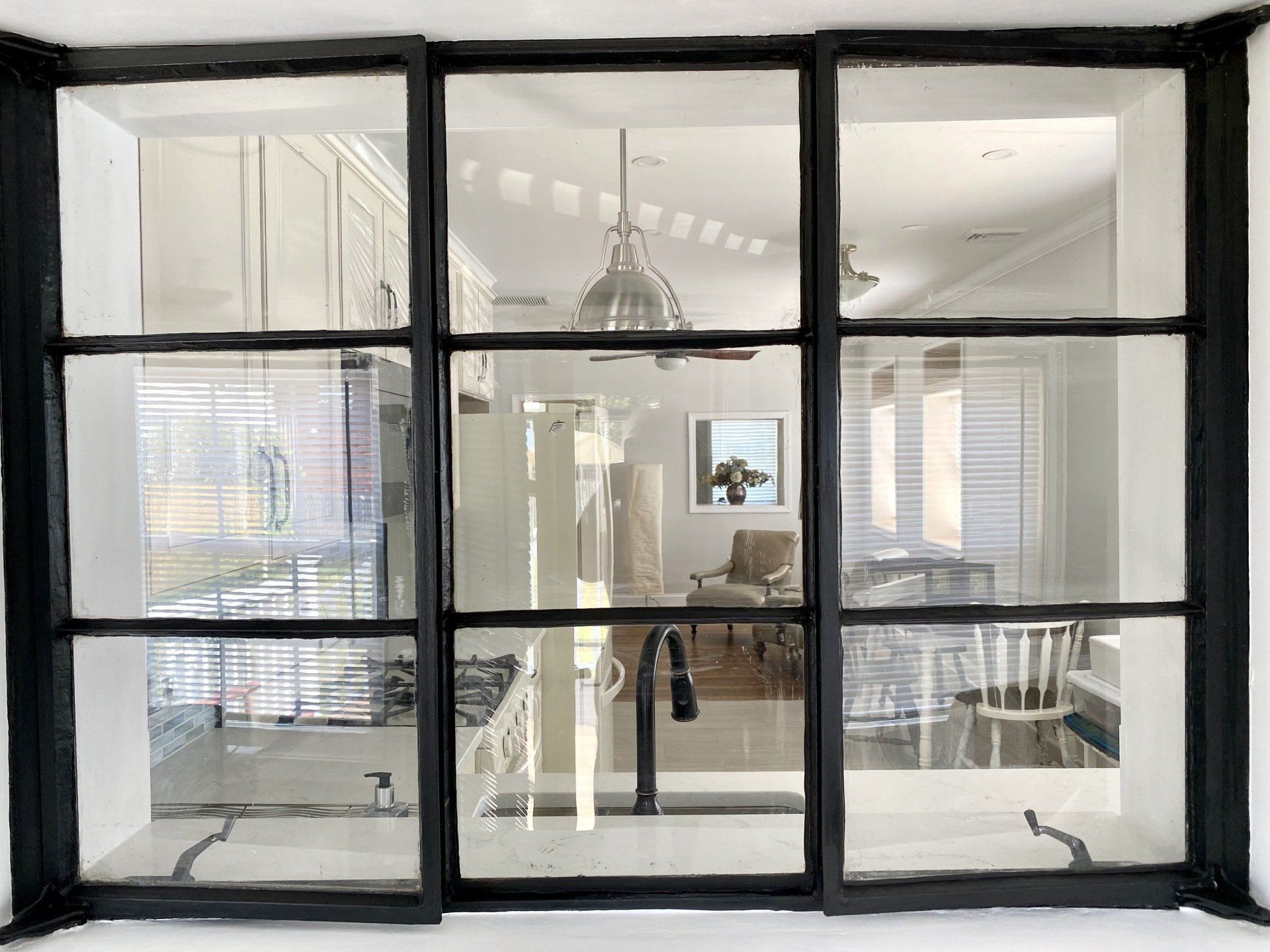 Black-framed window with six panes, looking into a kitchen with light fixtures, furniture, and white cabinetry.