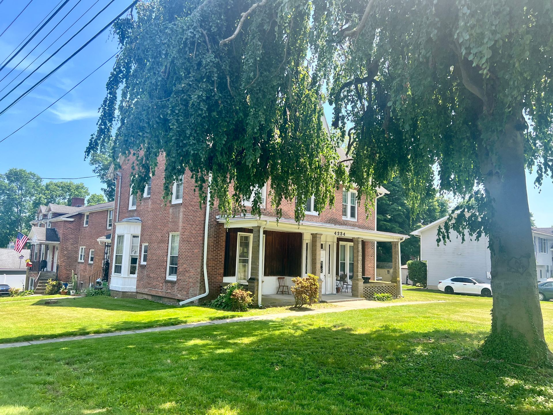 Brick house with porch, lush green lawn, partially shaded by large tree on a sunny day.