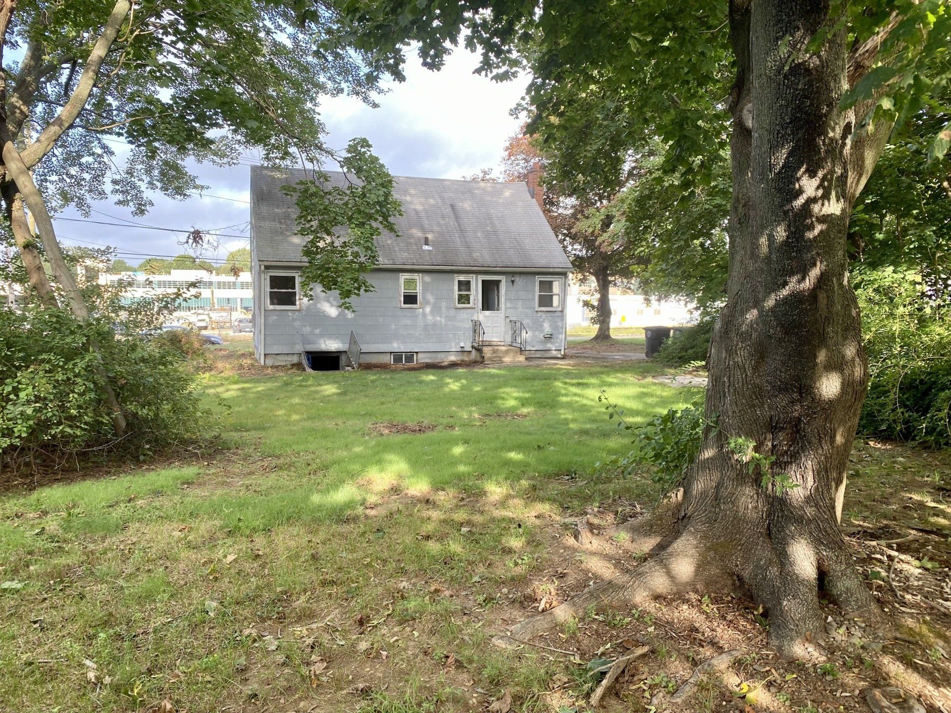 Backyard view of a small, gray house with trees and a grassy lawn on a sunny day.