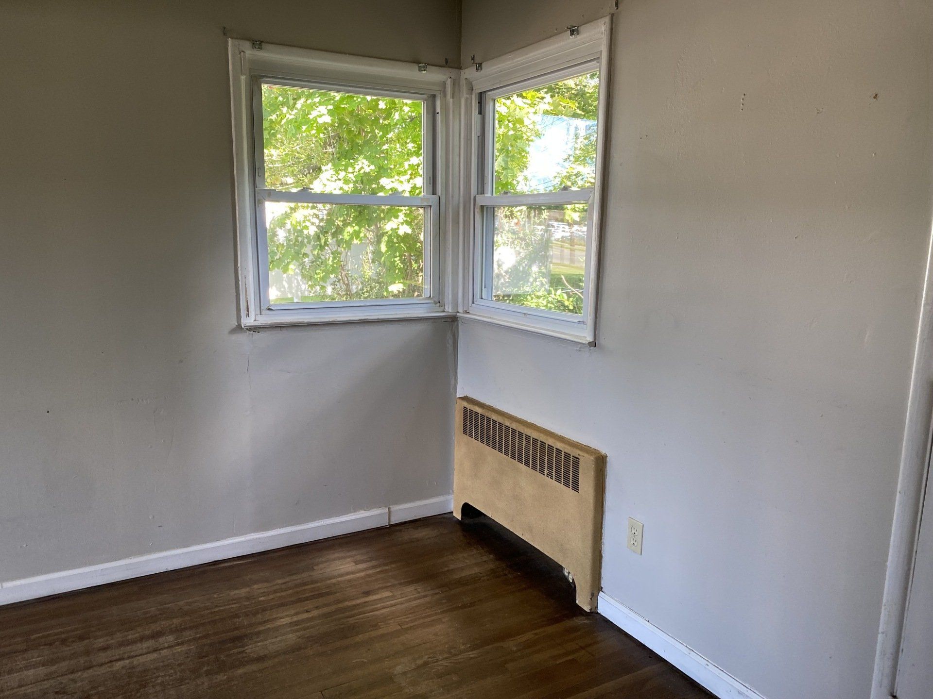 Empty room with corner windows and radiator, dark wood floor, light gray walls.
