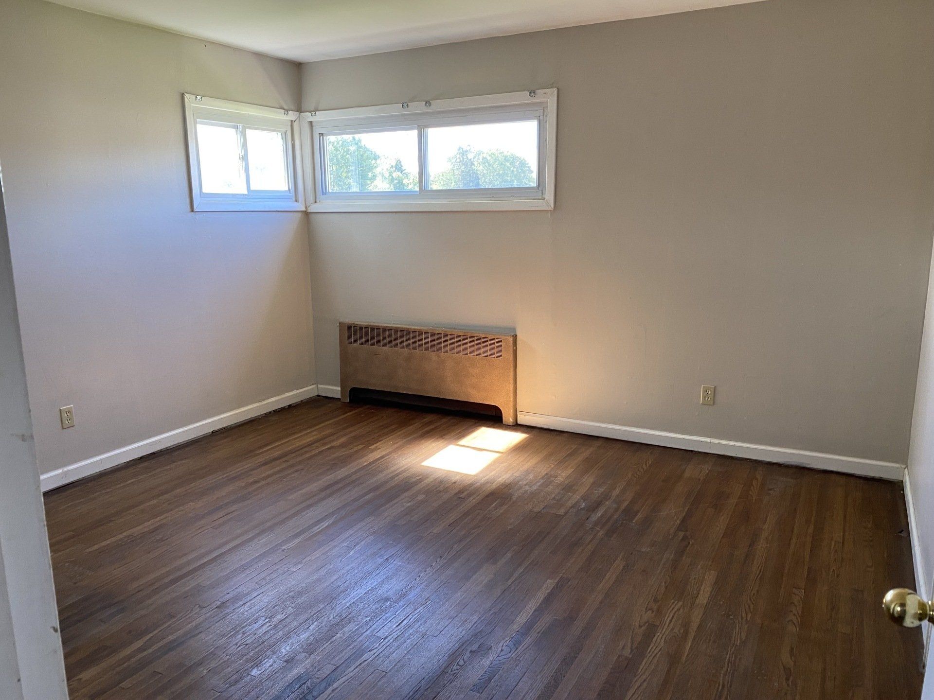Empty room with dark wood floors, radiator, and window. Beige walls.