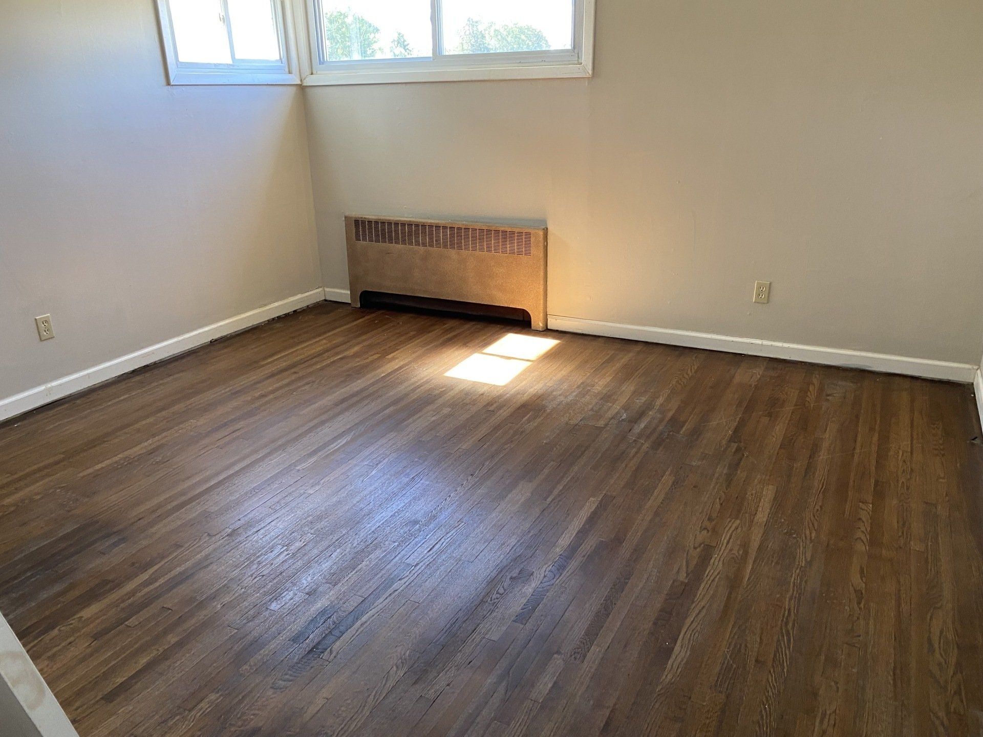 Empty room with hardwood floors, beige walls, and a radiator beneath a window.