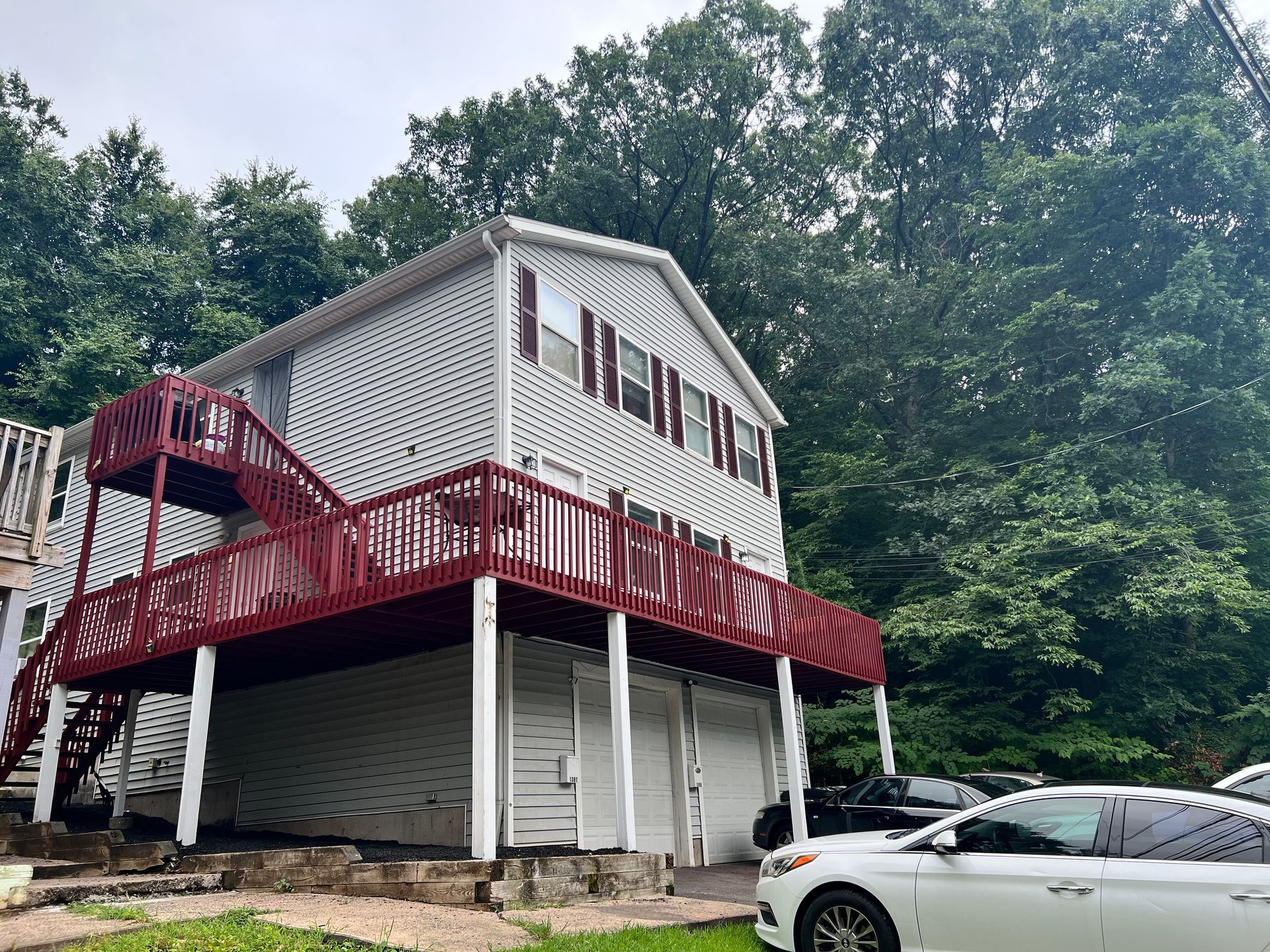 Two-story white house with red deck and stairs, set in front of green trees. A white car is parked nearby.