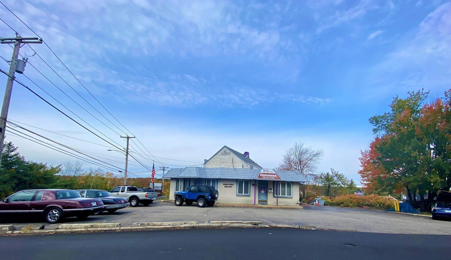 A small, single-story building with a parking lot and cars in front under a blue sky.