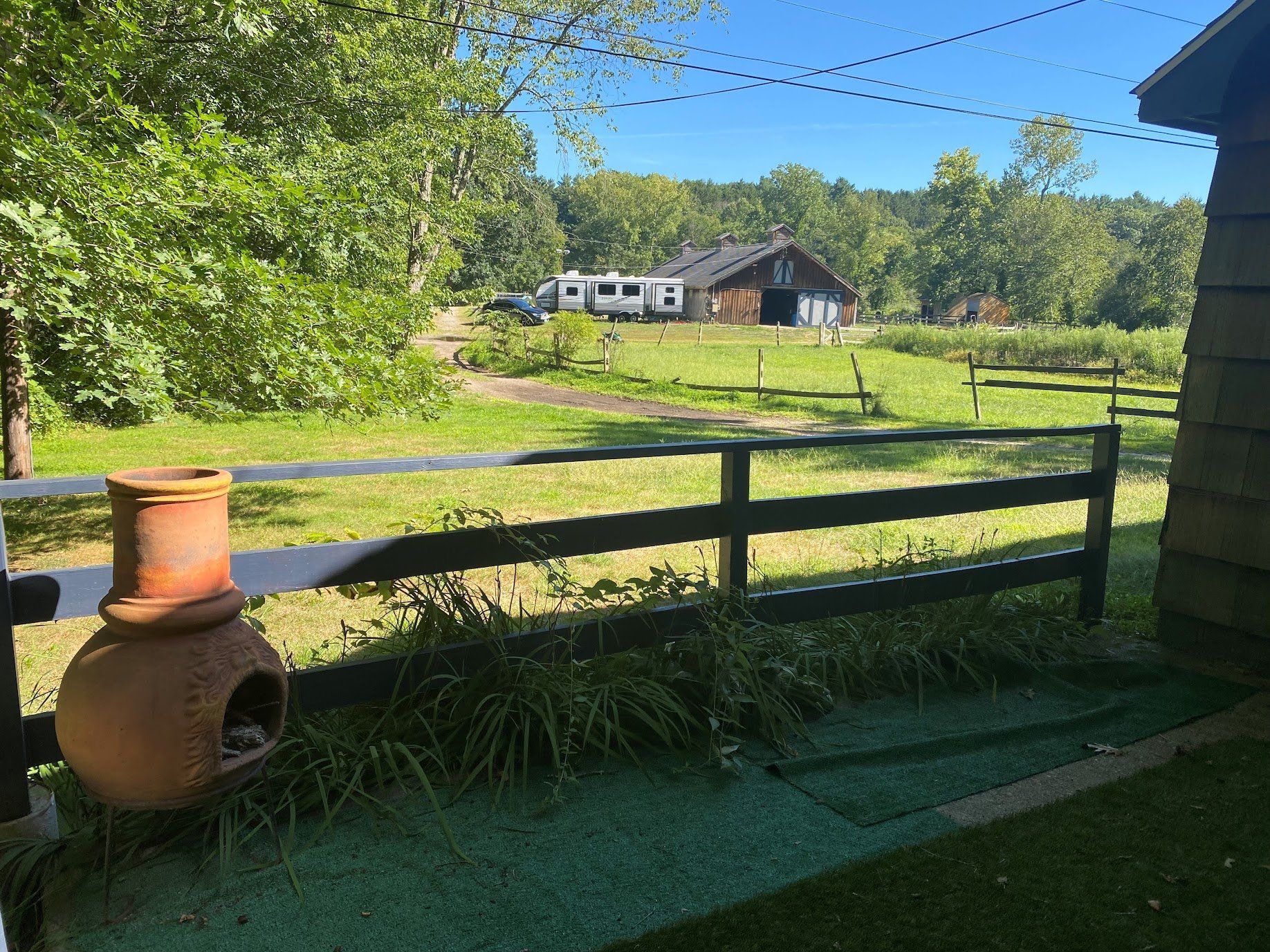View of a rural scene: grassy field, black fence, brown building, RVs, trees, and a terracotta chiminea.