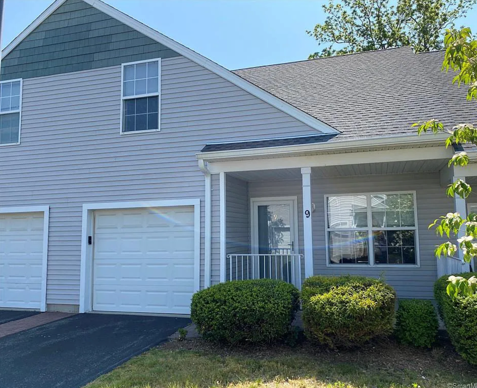 Gray townhouse with white garage door and porch, green bushes, and a blue sky.