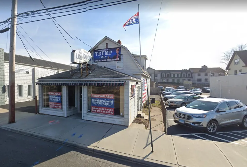 A liquor store with Trump signage and flags. Cars parked in adjacent lot, sunny day.