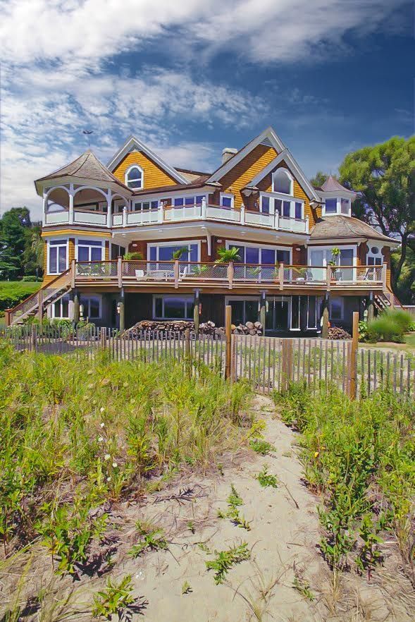 Large beach house, brown and yellow, with decks, set against a blue sky with clouds.