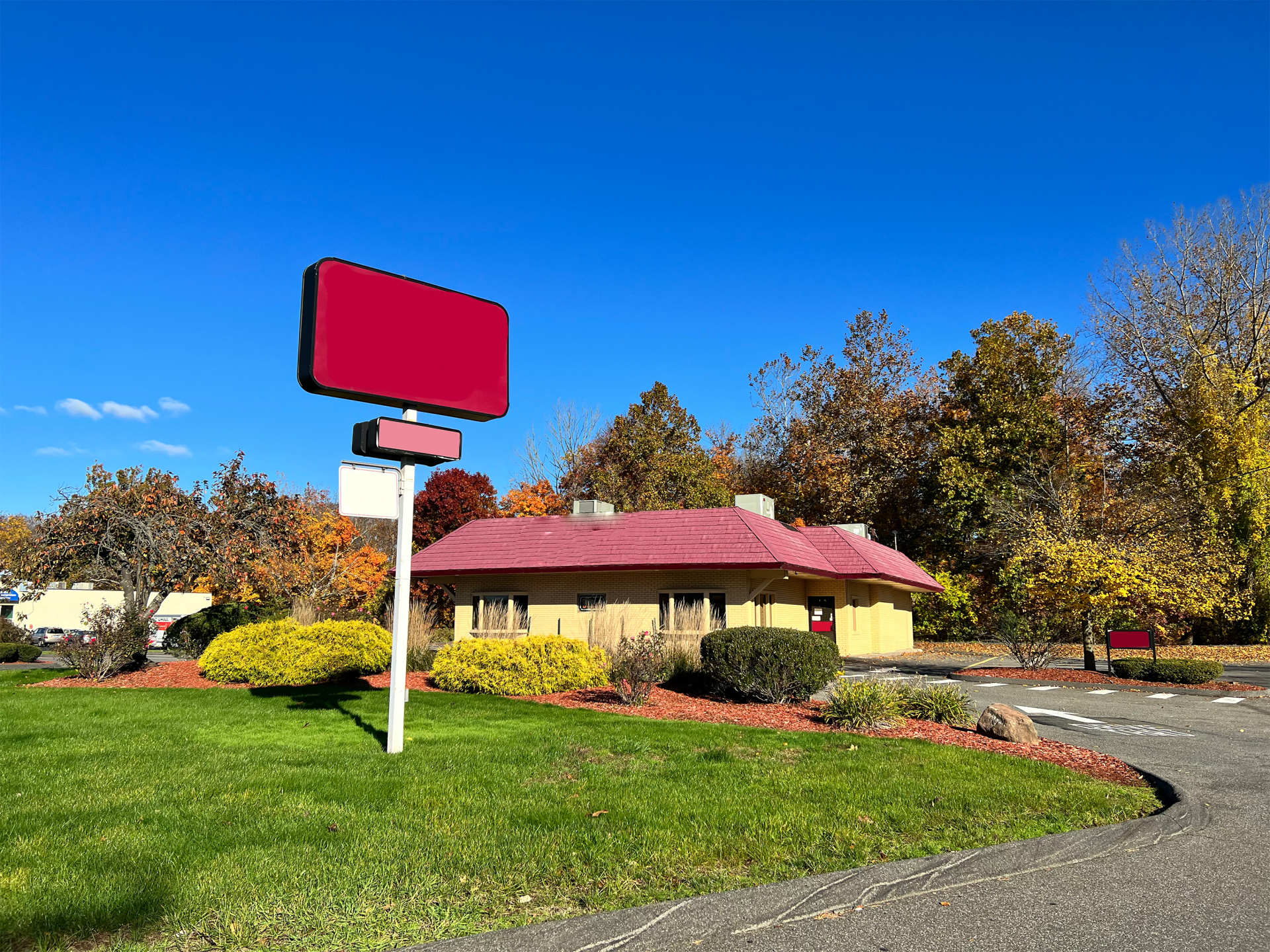 A small, yellow building with a red roof and blank red sign under a blue sky surrounded by trees and a green lawn.
