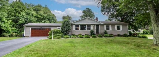 Gray house with a red garage door, surrounded by green grass and trees, under a blue sky.