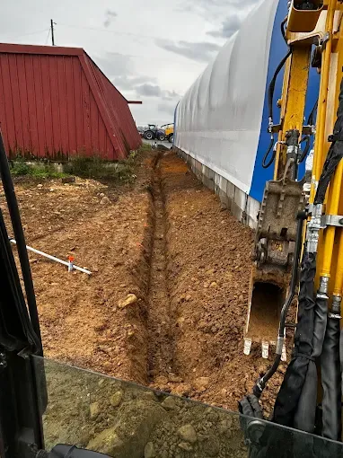 Narrow trench beside a white tarp-covered structure, with a red building and excavator arm on the right.