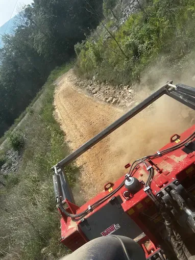 Red tractor mower on a winding dirt road beside dense green vegetation and hillside trees.