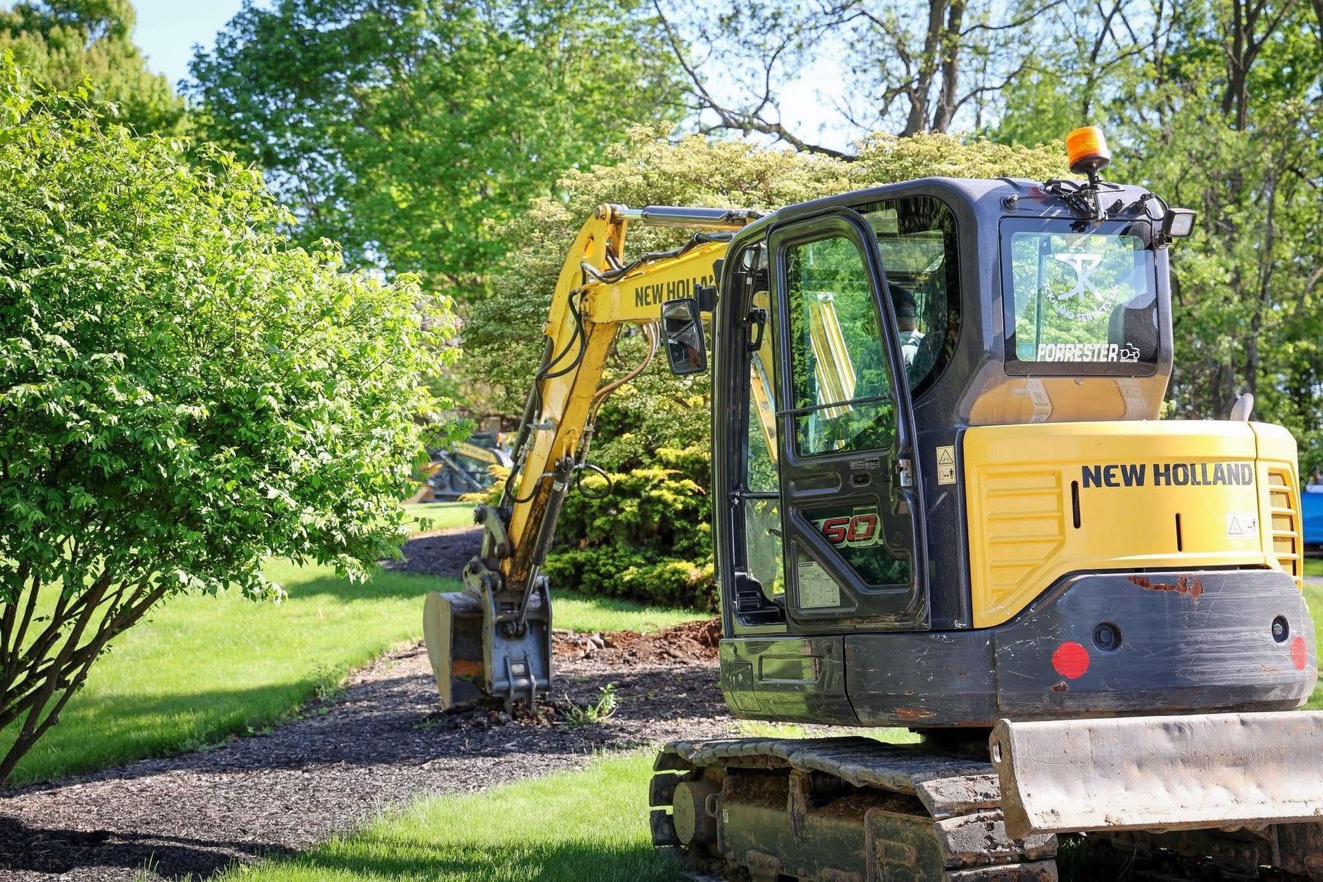 Yellow excavator clearing gravel beside a grassy path with trees and shrubs nearby