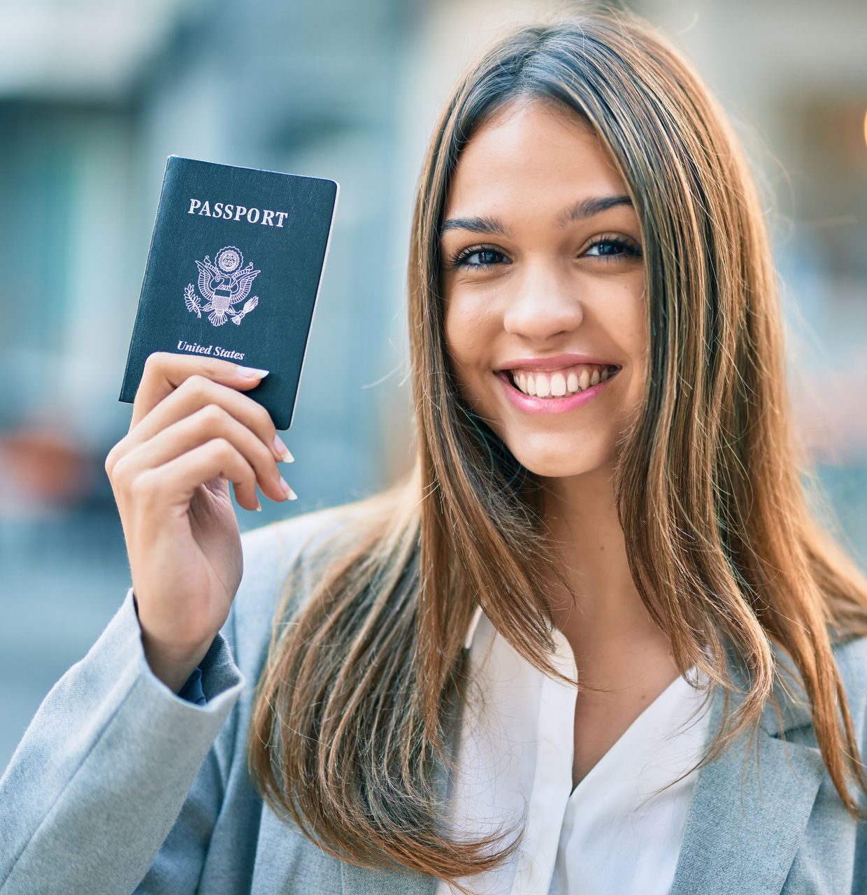 Smiling woman in a gray blazer holding up a passport outdoors