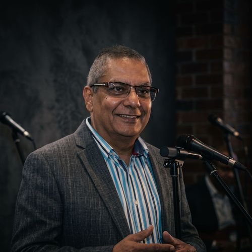 Studio headshot of a person wearing glasses, a purple shirt, and a dark blazer on a light background