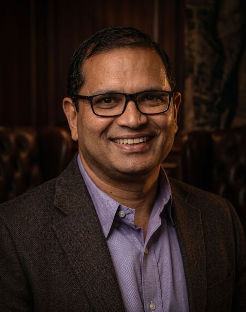 Studio headshot of a person wearing glasses, a purple shirt, and a dark blazer on a light background