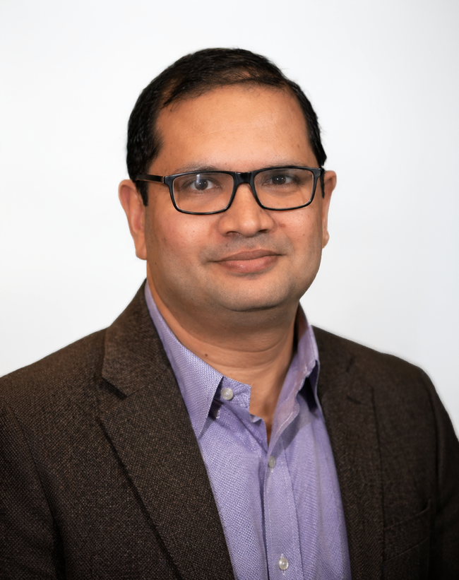 Studio headshot of a person wearing glasses, a purple shirt, and a dark blazer on a light background