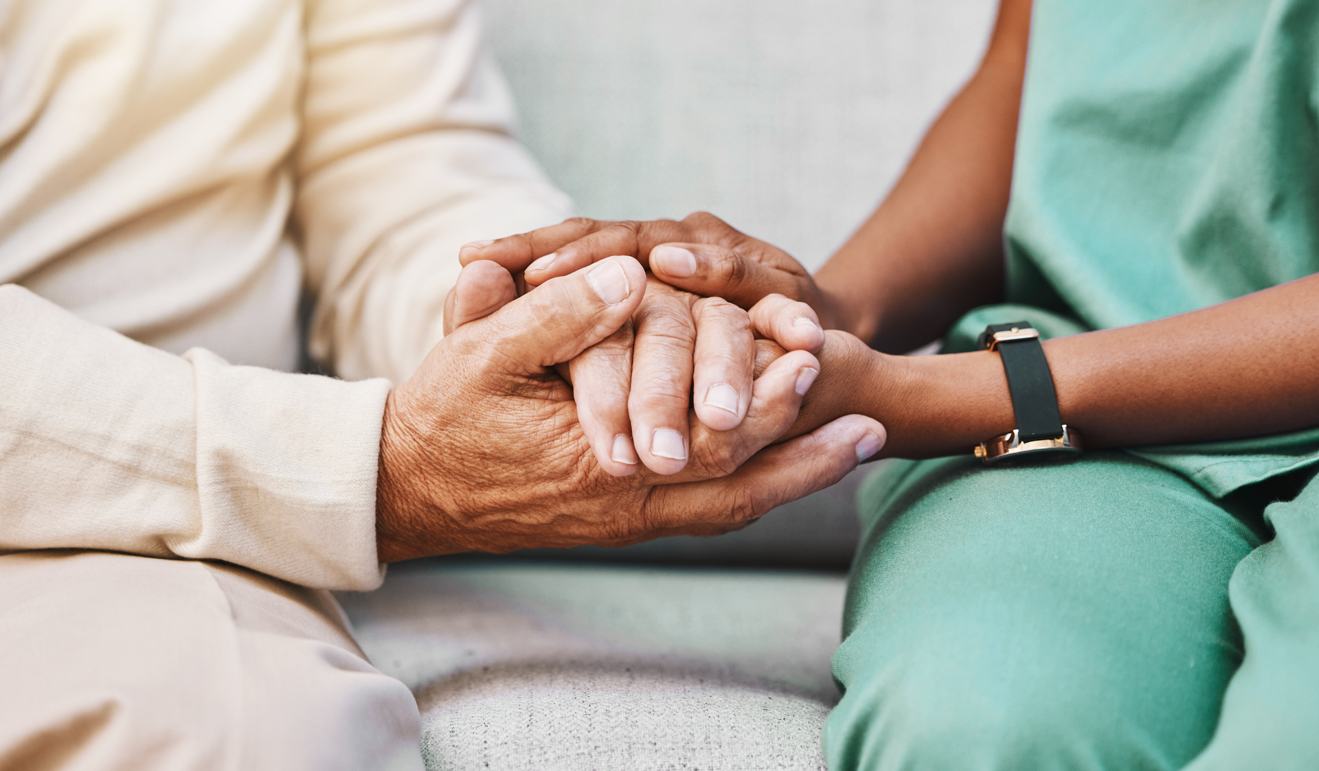 Two people holding hands on a couch, one in beige and one in green scrubs, with a wristwatch visibleS