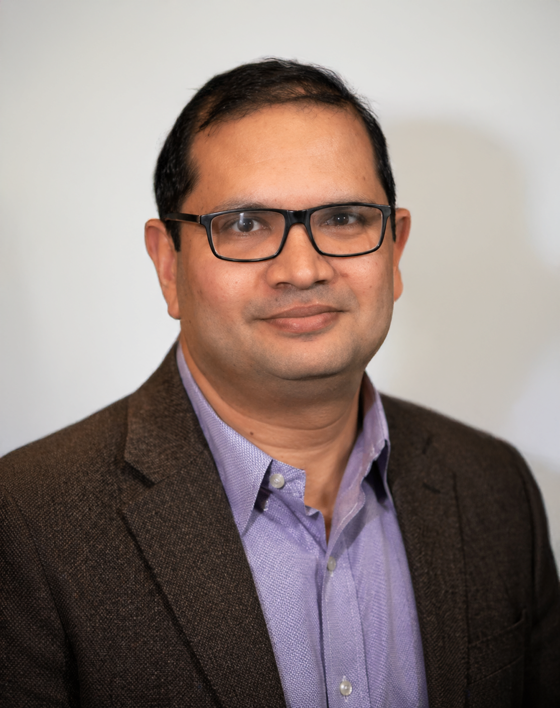 Portrait of a man wearing glasses, a brown blazer, and a light purple shirt against a plain background