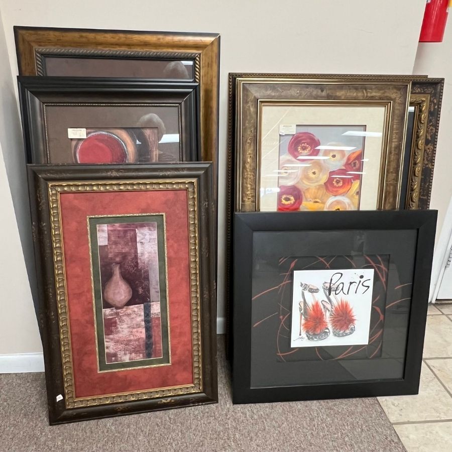 Five framed artworks leaning against a wall on a tiled floor, in brown, black, and gold frames.