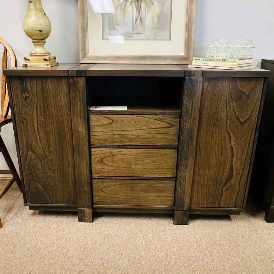 Dark wood sideboard with three drawers and cabinet doors, topped with a lamp and framed art.