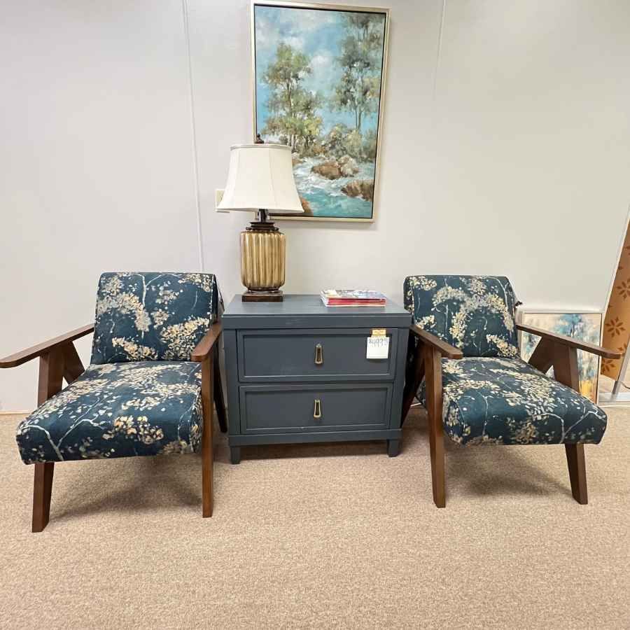 Two blue patterned armchairs flank a dark dresser with a lamp and coastal painting above it.