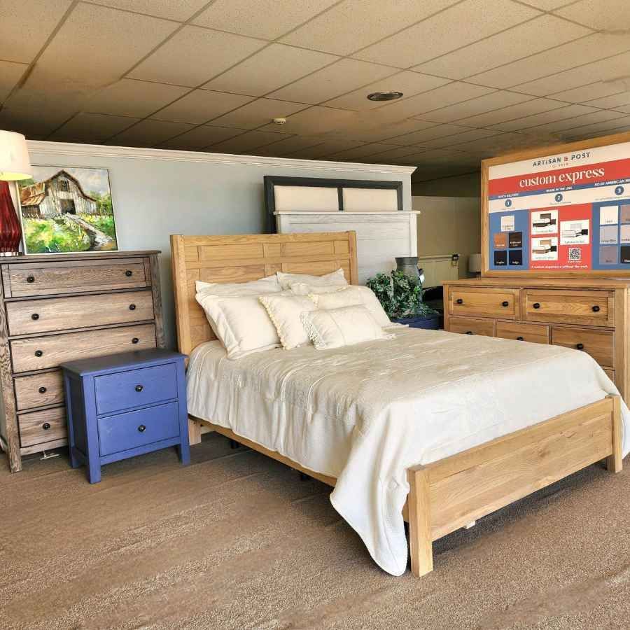 Bedroom furniture showroom with a light wood bed, dressers, and a blue nightstand on carpet.