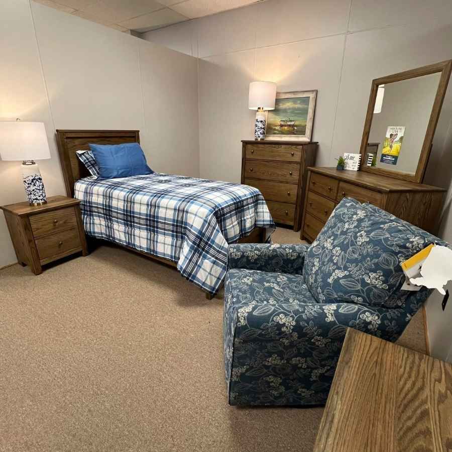 Small bedroom with a twin bed, blue armchair, dresser, lamp, and mirror on beige carpet.