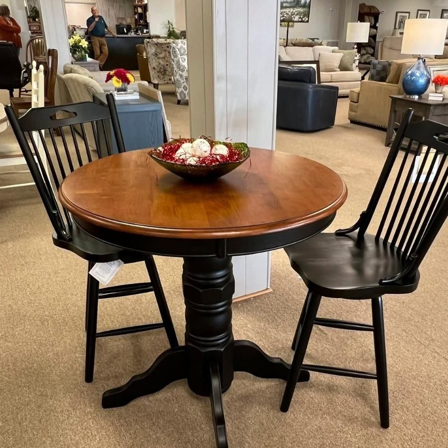 Round wooden table with two black chairs in a furniture showroom, with a decorative bowl on top.