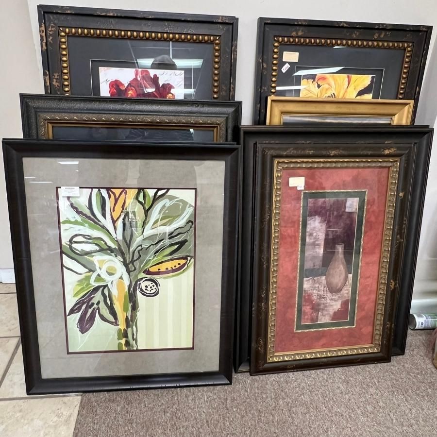 Assorted framed artwork leaning against a wall, including botanical and abstract prints.