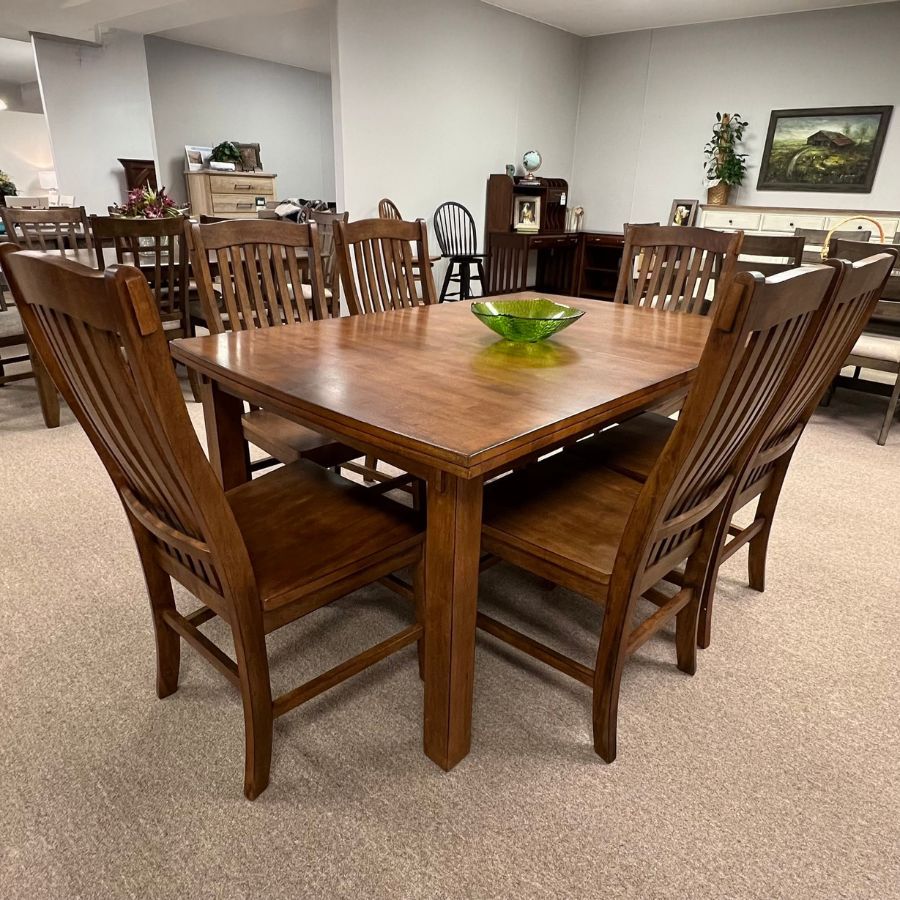 Wooden dining table and chairs in a showroom with beige carpet and white walls.