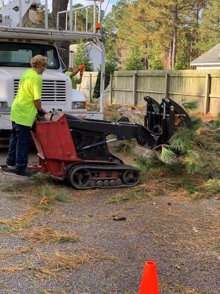 Worker beside tracked machine loading debris into a white truck in a yard with a wooden fence.