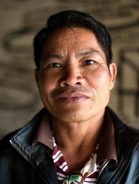 Close-up portrait of a man in a leather jacket and patterned shirt, standing against a blurred indoor background