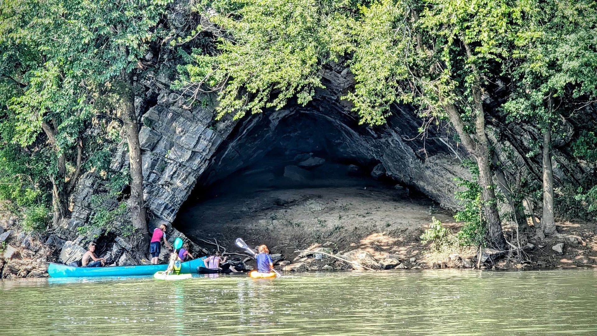 People in kayaks near a rocky cave entrance under dense green foliage by calm water