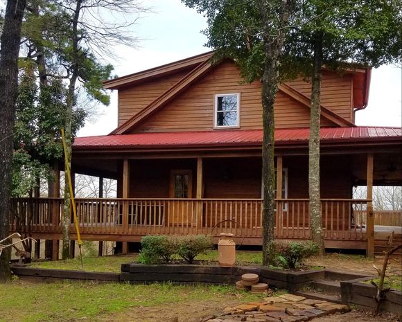 Wooden cabin with red roof and wraparound porch in a wooded yard