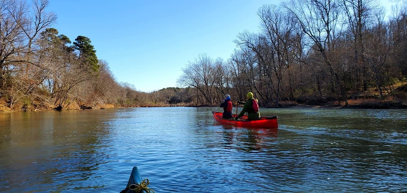 Two people paddling a red canoe on a calm river with trees along the banks under a clear blue sky