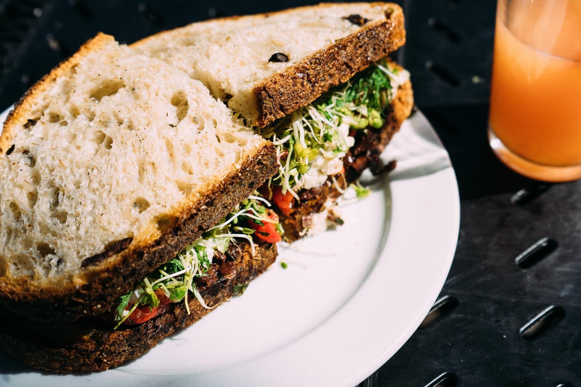 Large sandwich on a white plate with greens, beside a glass of orange drink